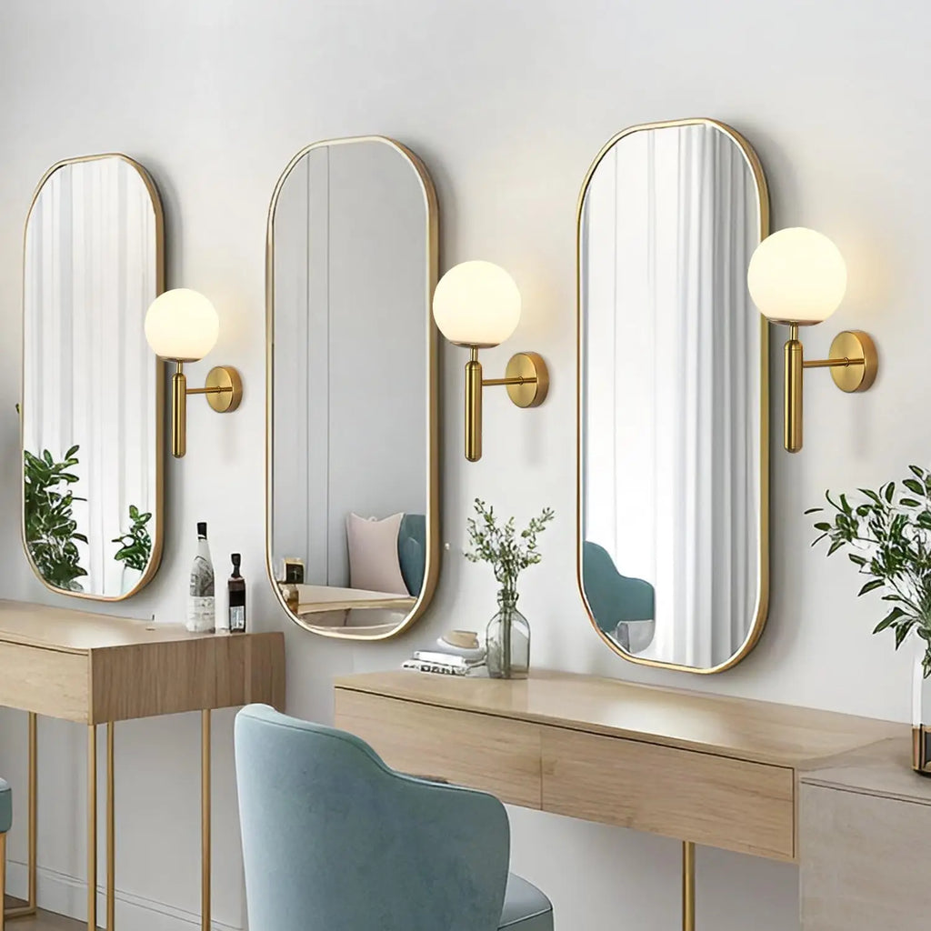 Trois appliques minimalistes dorées à globe de verre blanc, à côté de miroirs au-dessus d'une coiffeuse